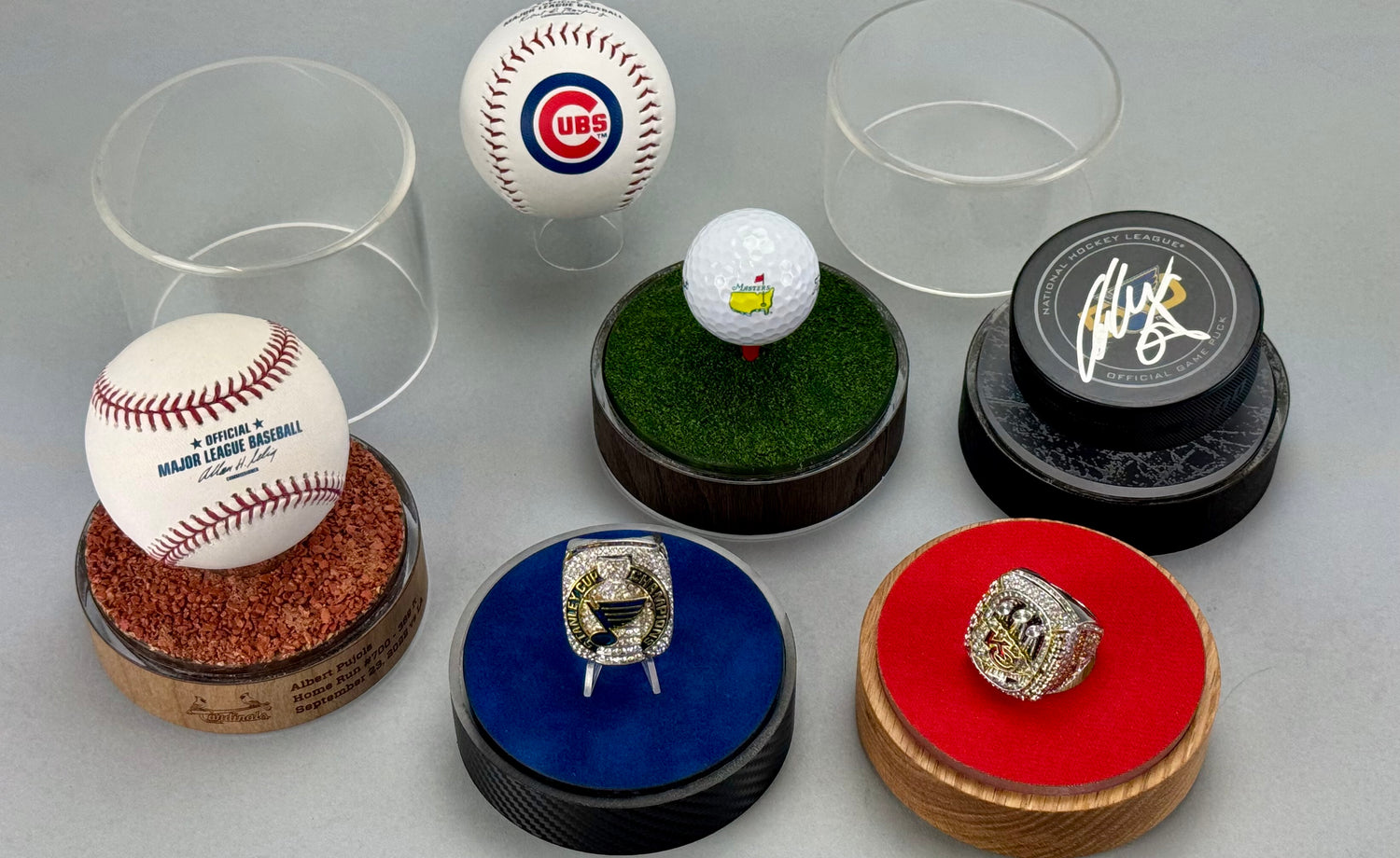 Five ProCase bases and two clear acrylic domes on a grey background. Each ProCase has a different base: light wood, brown wood, carbon fiber, and engraved Albert Pujols. They contain: Official MLB baseball, Cubs baseball, St. Louis Blues Stanley Cup ring, Kansas City Chiefs ring, Masters Tournament golfball, and a signed Alex Pietrangelo hockey puck. The ProCases display five different inserts: authentic infield clay, blue velvet, green putting, red cloth, and ice.